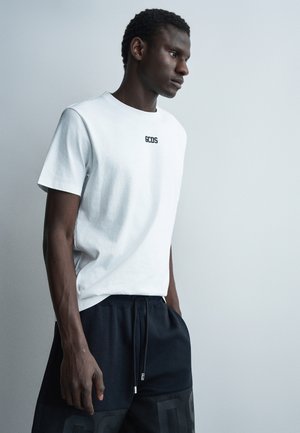 Νεαρός άνδρας που φοράει λευκό t-shirt GCDS και μαύρο σορτς, στέκεται μπροστά σε απλό ανοιχτό γκρι φόντο, κοιτάζοντας προς το πλάι με τα χέρια στις τσέπες.