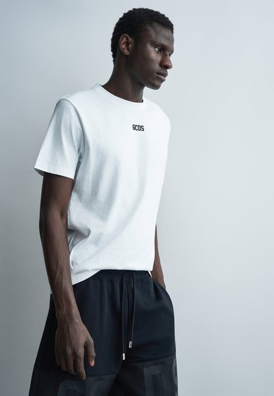 Jeune homme portant un t-shirt blanc GCDS et un short noir, debout devant un fond gris clair uni, regardant sur le côté avec les mains dans les poches.
