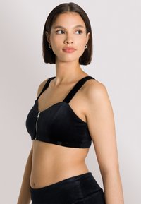 Reggiseno in corduroy nero con spalline larghe e zip frontale. Caratterizzato da un design strutturato e una superficie liscia. Abbinato a pantaloni neri.