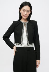 Μαύρο κοντό μπουφάν με γιακά, διακοσμημένο με ασημένιες χάντρες κατά μήκος των άκρων. Φοριέται πάνω από λευκό πουκάμισο και συνδυάζεται με μαύρο παντελόνι.