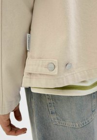 Beige denim jacket detail featuring silver snap buttons and a tag on the side, worn over a green-accented shirt and blue jeans.