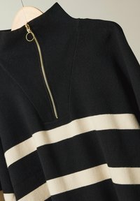 Schwarzer Strickpullover mit hohem Kragen, ausgestattet mit einem goldenen Reißverschluss und cremefarbenen horizontalen Streifen über dem Körper. Glatte Textur.