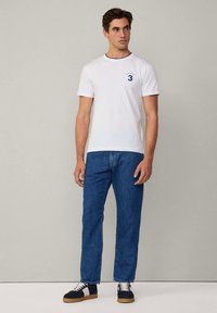Bílé bavlněné tričko s modrým límečkem a číslem "3" na přední straně, kombinované s modrými džínami a černými teniskami s bílými tkaničkami.