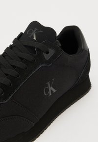 Svart sneaker med överdel i canvas och mocka, rund tå, snörning och präglad logotyp på sidan. Texturerad sula.