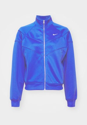 Blå zip-jakke med høy krage, laget av glatt stoff. Har en hvit Nike-logo på venstre bryst og ribbede mansjetter.