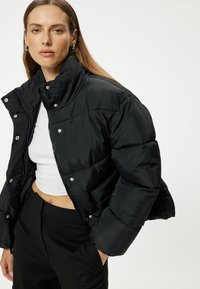 Chaqueta acolchada negra con cuello alto, cierre de cremallera y botones a presión. El material tiene una textura lisa y brillante con un patrón acolchado.