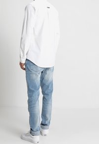 Witte lange mouw shirt met een achterpand, gecombineerd met lichtblauwe spijkerbroek en witte sneakers. Het shirt heeft een subtiele logopatch op de achterkant.