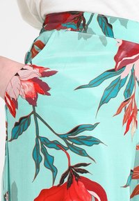 Spodnie w kwiatowy wzór mają miękki miętowy kolor z dużymi czerwonymi i różowymi kwiatami, gładką fakturę oraz boczne kieszenie.