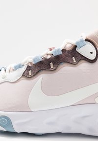 Ljusrosa sneakers med textilåverkan, vit swoosh-logotyp, metalliskt bronzfärgad snöröverdrag och blå accenter på snören och öglor.