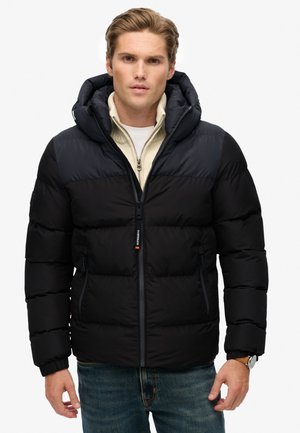 Un homme portant une veste matelassée noire et bleu marine par-dessus un pull zippé beige et un jean bleu, se tenant devant un fond blanc uni.