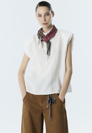 Femme portant un haut blanc sans manches aux épaules matelassées, une écharpe à motifs avec franges, et un pantalon marron à jambes larges avec cordons décoratifs.