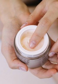 Szklany słoik z jasną śmietaną, biały korek, trzymany w dwóch rękach. Śmietana ma gładką, kremową konsystencję i jasny kolor.