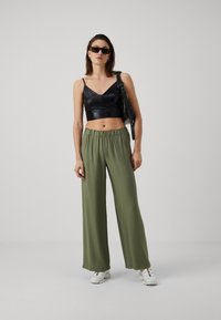 Fekete műbőr crop top, V-nyakkal, széles lábú olíva zöld nadrággal párosítva. A modellen fehér sportcipők és fekete napszemüveg van.