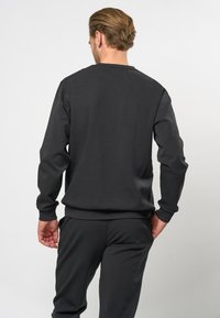 Muž s světlými hnědými vlasy, oblečený v jednoduché černé mikině s dlouhým rukávem a černých kalhotách, stojící zády k nám na bílém pozadí.