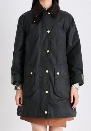 Personne portant un manteau noir boutonné avec deux grandes poches frontales et un col brun foncé, par-dessus une chemise à carreaux et une jupe marron.