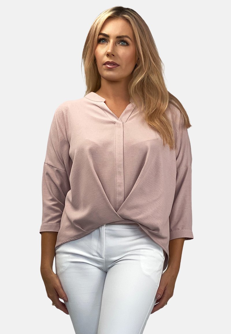 Jasnoróżowa bluzka w serek z rękawami 3/4, z drapowaną przednią częścią i miękkim materiałem. W połączeniu z białymi dopasowanymi spodniami.