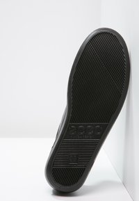 Svart gummisul med mönstrade spår för grepp, med "ECCO"-märket och en rektangulär logotyp, lutad mot en vit bakgrund.