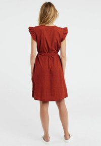 Červené šaty s volánovými rukávy, vypasovaným pasem a sukní ke kolenům. Mají tečkovaný vzor a texturovanou látku. Zobrazeno ze zadní strany.