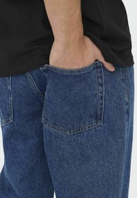 Plave traperice s relaxed fit krojem, s stražnjim džepom i kontrastnim šavovima. Ruka je odložena u prednjem džepu.