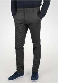 Pantaloni slim-fit grigio scuro realizzati in tessuto misto cotone, con chiusura a bottone, tasche e dettagli testurizzati sottili. Indossati con sneakers blu navy.