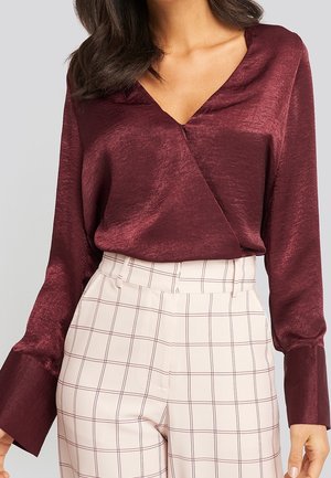 Mujer con una blusa de satén granate de manga larga y escote en V metida en pantalones crema de cintura alta con un patrón de cuadros marrón.