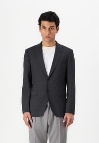 Mörkgrå blazer med två framknappar, slagkragar och en bröstficka. Buren över en ljusfärgad t-shirt och kombinerad med randiga byxor.