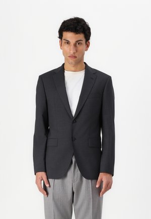 Dunkelgrauer Blazer mit zwei Frontknöpfen, Reverskragen und einer Brusttasche. Getragen über einem hellen T-Shirt und kombiniert mit gestreiften Hosen.
