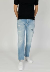 Vaqueros rasgados de color azul claro con detalles rotos, corte recto y costuras en contraste, combinados con zapatillas blancas.