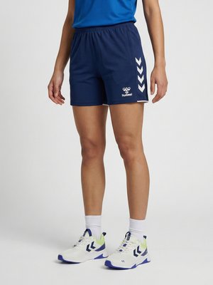 Osoba nosząca granatowe krótkie spodenki sportowe z białymi logo w kształcie szewronu, białe skarpety i białe oraz niebieskie buty sportowe na szarym tle.