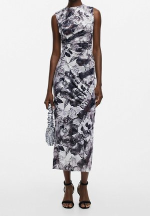 Robe midi slim sans manches avec motif floral noir et blanc, détails froncés, associée à des talons noirs à bride cheville et un sac à main froncé argenté.
