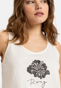 White tank top featuring a textured black hibiscus flower graphic and the word "Roxy" underneath in a handwritten style.