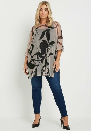 Durchsichtige graue Bluse mit schwarzem Blumenmuster, weiten Ärmeln und seitlichen Schlitzen, kombiniert mit dunkelblauen Jeans und schwarzen High Heels.