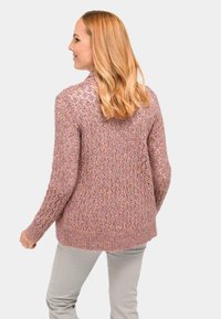 Rosa gestrickte Strickjacke mit lockerem Gewebe und Rautenmuster, ausgestattet mit einem hohen Kragen und geripptem Saum. Getragen mit hellen Hosen.