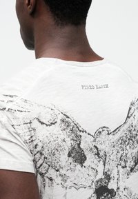 T-shirt branca com um grande gráfico preto de asas de anjo nas costas. O tecido tem uma aparência texturizada, enfatizando um estilo casual.