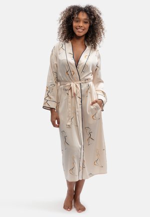 Woman with curly hair smiling, wearing light-colored patterned silk robe tied at the waist, standing barefoot against a plain background.