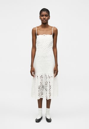 Woman wearing a white sleeveless midi dress with eyelet cutouts, bow-tied shoulder straps, white socks, and black flat shoes, standing on white background.