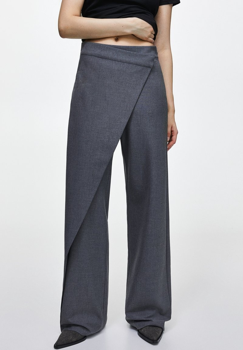 Personne portant un pantalon large gris foncé taille haute avec un panneau avant superposé et des chaussures noires pointues, debout devant un fond uni.