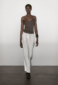 Bruine tanktop en witte linnen broek. Het model draagt zwarte sandalen en zilveren armbanden. Eenvoudig, minimalistisch ontwerp tegen een neutrale achtergrond.