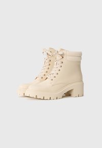 Bottines à talons hauts beiges en matériau synthétique lisse, avec un col côtelé, un laçage à l'avant et une semelle en caoutchouc épaisse et crantée.