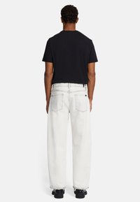 Maglietta nera a maniche corte, vestibilità rilassata, abbinata a jeans bianchi a gamba larga con orli sfrangiati. Scarpe nere completano il look.