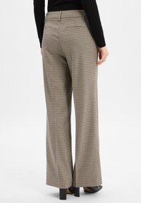 Frau trägt beiger und schwarzer, hochtaillierter, weiter Beinkleid mit Karomuster, kombiniert mit schwarzen High Heels und einer figurbetonten schwarzen Bluse.