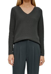 Frau in einem dunkelgrauen V-Ausschnitt-Rippstrickpullover, der in hoch taillierte, gefältelte dunkelgraue Hose gesteckt ist, steht mit entspannten Armen.