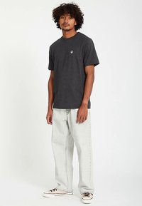 Graues T-Shirt mit kurzen Ärmeln und rundem Ausschnitt sowie kleinem Logo auf der linken Brust, kombiniert mit hellgrauen Jeans und schwarz-cremefarbenen Sneakers.