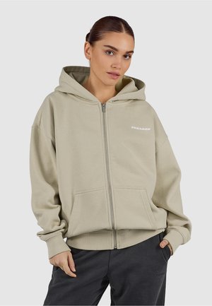 Kobieta ubrana w beżową bluzę z kapturem na zamek błyskawiczny i przednimi kieszeniami, w ciemnych spodniach, stojąca z jedną ręką w kieszeni.