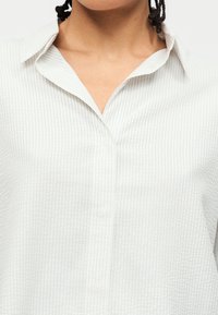 Άτομο που φοράει λευκό πουκάμισο με κάθετες ρίγες και κουμπιά, με ανοιχτό γιακά και πλεξούδα που φαίνεται στο πάνω μέρος.