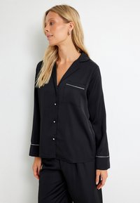 Zwarte satijnen pyjama set met lange mouwen, een zak en zilveren bies langs de manchetten en de kraag. Voorzien van parelachtige knoopjes aan de voorkant.