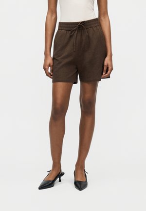 Personne portant un short à cordon de serrage marron foncé, un haut blanc sans manches et des chaussures à talons bas noires à bout pointu, debout devant un fond blanc.