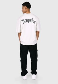 Hvit oversized t-skjorte med svart gotisk tekst "Dropsize" på baksiden, kombinert med svarte cargo-bukser og hvite sneakers.