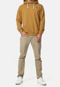 Sennepsfarvet hoodie med hvide snore og logo på brystet, sammen med lyse beige bukser og hvide sneakers. Glat stoftekstur.
