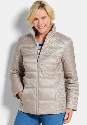 Donna sorridente, indossa una giacca trapuntata beige chiaro sopra una camicia a righe, con le mani nelle tasche, in piedi contro uno sfondo bianco.